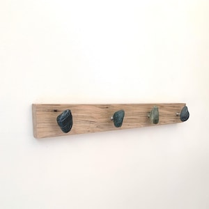 May include: A wooden coat rack with four stone-shaped knobs. The wood has a natural grain and a light brown color. The knobs are various shades of gray and green. The rack is mounted on a white wall.