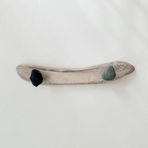 May include: A horizontal piece of light brown driftwood featuring two attached stones. One stone is solid black and positioned on the left side, while the other stone is a muted green color and is positioned on the right side of the wood.