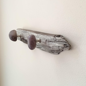 May include: A wall-mounted coat rack featuring a piece of weathered gray driftwood with a rough, textured surface. Two smooth, rounded, purple-toned stone knobs are attached to the wood with silver-colored metal hardware for hanging items.