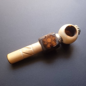 Può includere: Una pipa da fumo in legno con una ciotola bianca e un gambo in legno marrone. Il gambo ha un design intagliato e la ciotola ha un piccolo design a fiore.