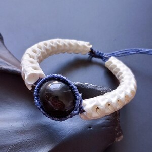 Peut inclure: Un bracelet artisanal avec une pierre noire polie au centre, entourée d'un cordon bleu tressé. Le bracelet est composé de segments blancs en forme d'os. Sur un fond sombre et texturé.