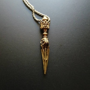 Könnte beinhalten: Eine detaillierte Anhängerkette mit einem goldfarbenen, verzierten, dolchförmigen Anhänger mit detailliertem Gesichtsdesign. Der Anhänger hängt an einer silberfarbenen Kette. Der Hintergrund ist eine dunkle, einfarbige Farbe.