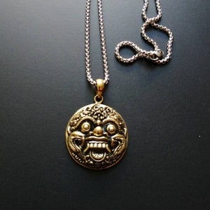 May include: A gold-toned pendant necklace featuring an intricate, ornate design of a mythical creature's face. The pendant is attached to a silver chain.
