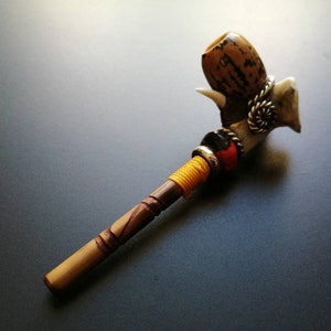 May include: A wooden smoking pipe with a decorative bowl made of natural materials. The bowl is decorated with a spiral design and a small, white horn. The pipe is wrapped with yellow and red string.