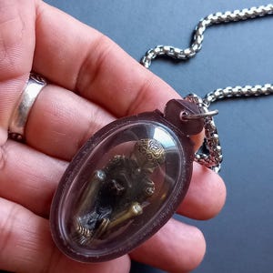 Puede incluir: Un collar con colgante que presenta una pequeña y detallada figura encerrada en un colgante de plástico transparente de forma ovalada. La figura es oscura con detalles dorados, y el colgante cuelga de una cadena plateada. El fondo es gris oscuro liso.