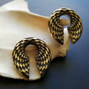 May include: Two gold-coloured metal ear gauges with a geometric pattern. The gauges are shaped like a horseshoe with a pointed end.