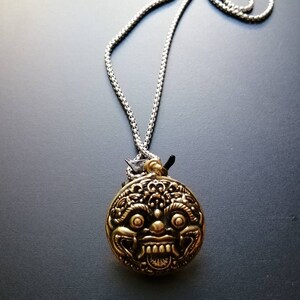 K&ouml;nnte beinhalten: Eine silberne Kettenkette mit einem runden Anh&auml;nger, der ein detailliertes goldfarbenes Gesicht mit einem grimmigen Ausdruck zeigt.