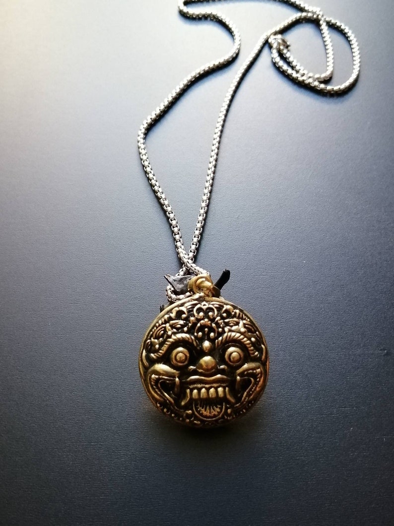 K&ouml;nnte beinhalten: Eine silberne Kettenkette mit einem runden Anh&auml;nger, der ein detailliertes goldfarbenes Gesicht mit einem grimmigen Ausdruck zeigt.