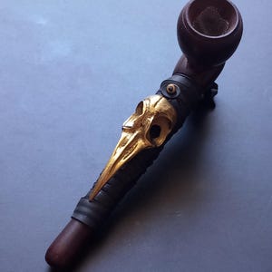 Op de afbeelding: Een donkerbruine houten rookpijp met een metallic goudkleurig vogel-schedelornament bevestigd aan de steel. De pijp heeft een ronde kop met een metalen gaasje aan de binnenkant en een glad, cilindervormig mondstuk aan het uiteinde.