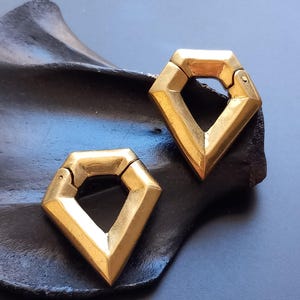 May include: Two gold-colored, diamond-shaped earrings with hinged clasps. The earrings have a geometric design with sharp angles and a polished finish. They are displayed on a dark, textured surface.