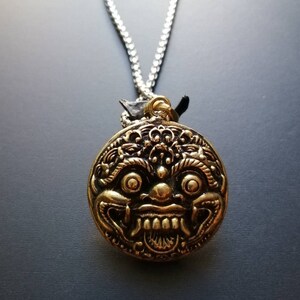 K&ouml;nnte beinhalten: Eine goldfarbene Halskette mit einem runden Anh&auml;nger, der ein detailliertes, stilisiertes Gesicht mit gro&szlig;en Augen und einem breiten Mund zeigt. Der Anh&auml;nger ist an einer silbernen Kette befestigt.