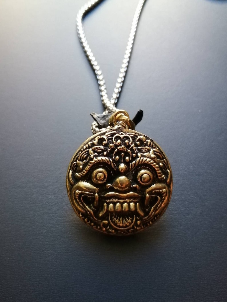 K&ouml;nnte beinhalten: Eine goldfarbene Halskette mit einem runden Anh&auml;nger, der ein detailliertes, stilisiertes Gesicht mit gro&szlig;en Augen und einem breiten Mund zeigt. Der Anh&auml;nger ist an einer silbernen Kette befestigt.