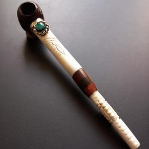 May include: A handcrafted tobacco pipe. The pipe has a dark brown bowl and a cream-coloured stem with carved leaf designs and a brown leather band. A green stone is set in a silver-coloured ring near the bowl.