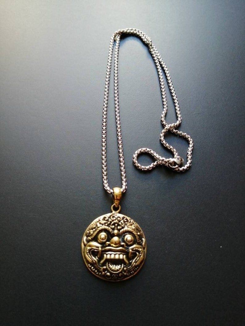 K&ouml;nnte beinhalten: Eine goldfarbene Halskette mit einem detaillierten, verzierten Design eines mythischen Gesichts. Der Anh&auml;nger ist an einer silbernen Kette befestigt.