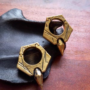 Op de afbeelding: Een paar oorhangers in messingkleur met een zeshoekig frame, een getextureerd en verweerd oppervlak en een centrale ronde opening. Elk sieraad is voorzien van een puntige, naar beneden gerichte stekel aan de onderkant met een metalen afwerking.