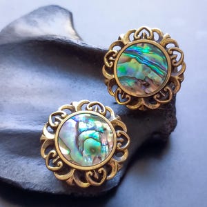 May include: A pair of gold earrings with a round abalone shell inlay. The earrings have a delicate, ornate design.