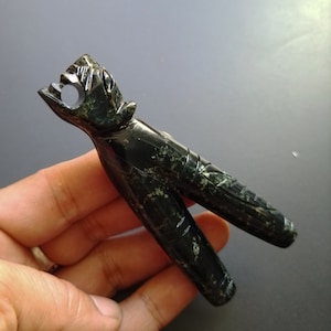 Op de afbeelding: Een donkergroene jade-carving van een gestileerd menselijk figuur met een vogelachtig hoofd. De figuur heeft twee benen en een gat in het hoofd.