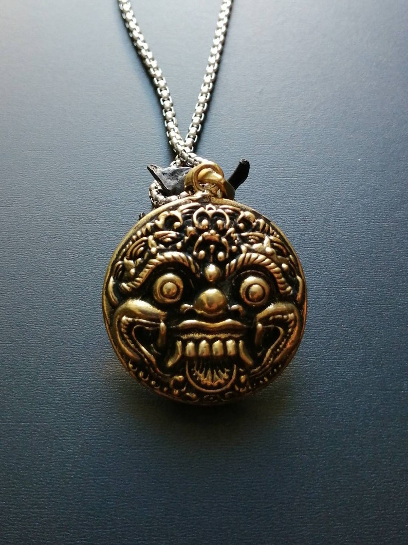 K&ouml;nnte beinhalten: Eine silberne Kettenkette mit einem runden goldenen Anh&auml;nger, der ein detailliertes, stilisiertes Gesicht mit einem zahnigen Grinsen zeigt.