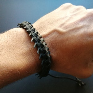 Peut inclure: Un bracelet en caoutchouc noir avec un motif texturé en forme d'épine dorsale. Le bracelet est réglable avec un cordon noir et un nœud.