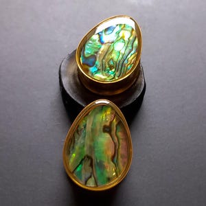 May include: Two gold-toned ear gauges with a green, blue, and iridescent abalone shell inlay. The gauges are oval shaped and have a smooth, polished finish.