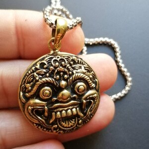 K&ouml;nnte beinhalten: Eine goldfarbene Halskette mit einem runden, detaillierten, verzierten Anh&auml;nger, der das Gesicht einer mythischen Kreatur darstellt. Der Anh&auml;nger ist an einer silbernen Kette befestigt.