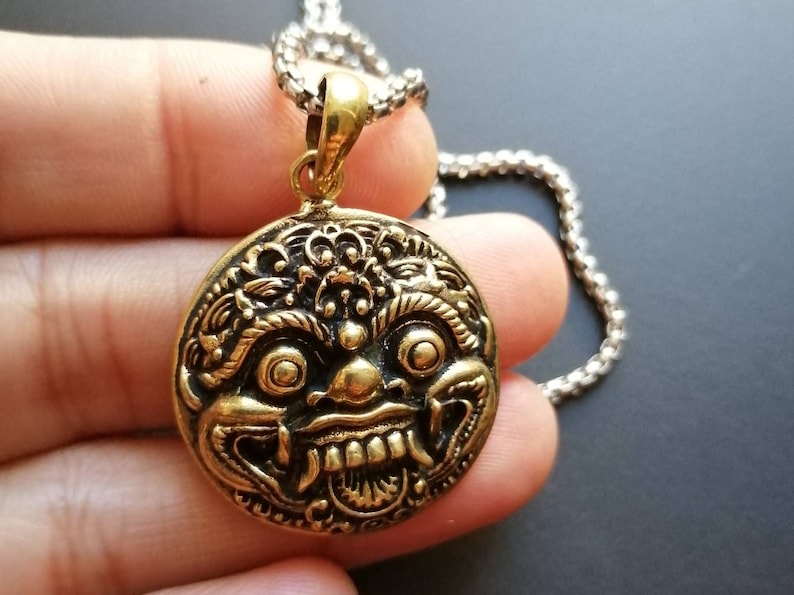 K&ouml;nnte beinhalten: Eine goldfarbene Halskette mit einem runden, detaillierten, verzierten Anh&auml;nger, der das Gesicht einer mythischen Kreatur darstellt. Der Anh&auml;nger ist an einer silbernen Kette befestigt.