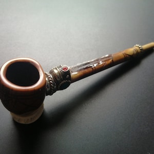May include: A brown wooden pipe with a decorative silver band and a red and blue gemstone. The pipe has a bowl with a brown and white pattern.