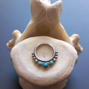 Può includere: Un anello per il setto nasale in argento con pietre turchesi. L'anello presenta un design a cerniera ed è decorato con piccole perle d'argento e tre pietre turchesi più grandi. Il gioiello è esposto su un oggetto color osso su sfondo blu.