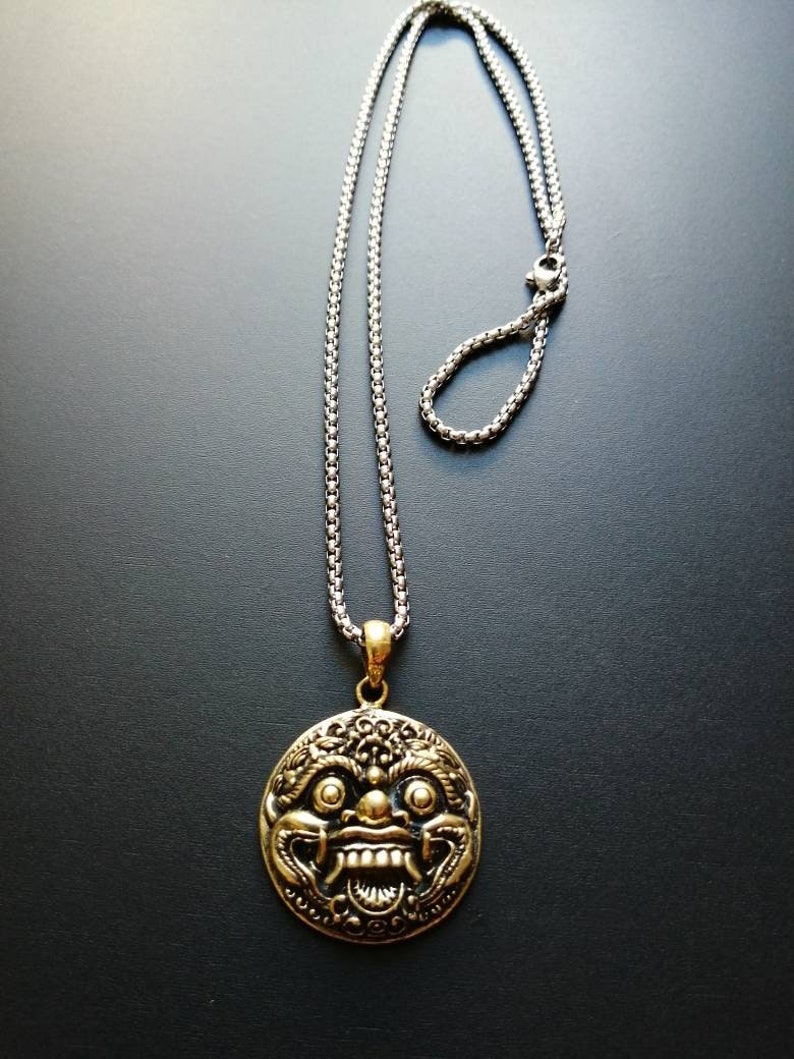 K&ouml;nnte beinhalten: Eine silberne Kettenkette mit einem goldenen Anh&auml;nger, der ein detailliertes, stilisiertes Gesicht mit einem gro&szlig;en Mund und Z&auml;hnen zeigt. Der Anh&auml;nger ist oval.