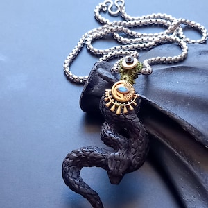 Op de afbeelding: Een zwarte slangen hanger met getextureerde schubben, hangend aan een zilverkleurige boxketting. De slang is voorzien van een goudkleurige zon-bedel met een centrale blauwe steen, bevestigd door een groene koordknoop met een kleine zwarte kraal aan de bovenkant.