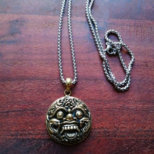 K&ouml;nnte beinhalten: Eine goldfarbene Halskette mit einem runden Anh&auml;nger, der ein detailliertes Design eines mythischen Gesichts zeigt. Die Kette ist aus Silber.