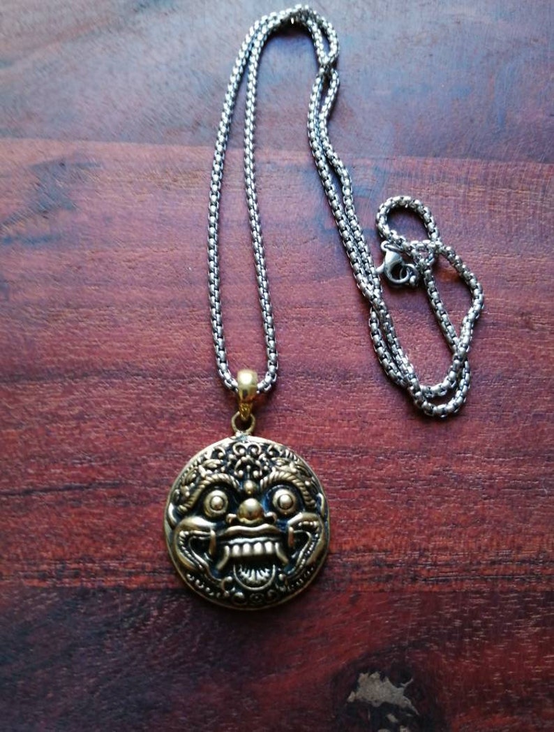 K&ouml;nnte beinhalten: Eine goldfarbene Halskette mit einem runden Anh&auml;nger, der ein detailliertes Design eines mythischen Gesichts zeigt. Die Kette ist aus Silber.