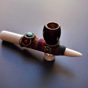 May include: A handcrafted tobacco pipe with a white mouthpiece and a dark brown bowl. The pipe features decorative metal accents, a turquoise stone, and colorful woven fabric. The pipe is approximately 15 cm long.