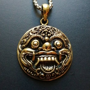 K&ouml;nnte beinhalten: Eine goldfarbene Halskette mit einem runden Anh&auml;nger, der ein detailliertes, verziertes Design eines mythischen Gesichts mit gro&szlig;en Augen, einem breiten Mund und scharfen Z&auml;hnen zeigt.