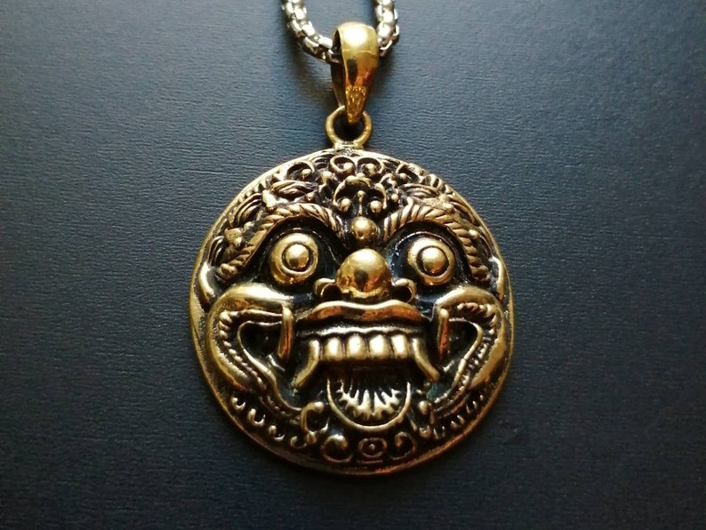K&ouml;nnte beinhalten: Eine goldfarbene Halskette mit einem runden Anh&auml;nger, der ein detailliertes, verziertes Design eines mythischen Gesichts mit gro&szlig;en Augen, einem breiten Mund und scharfen Z&auml;hnen zeigt.