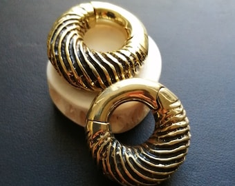 Puede incluir: Dos medidores de oreja de metal de color dorado con un diseño en espiral. Los medidores están sobre una superficie blanca.