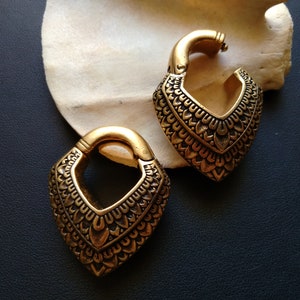 May include: A pair of gold-coloured earrings with a heart-shaped design. The earrings feature a detailed, textured pattern and a hinged clasp. They are displayed on a white, organic-shaped surface against a dark background.