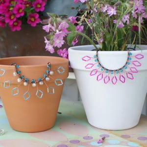 Könnte beinhalten: Zwei Terrakotta-Blumentöpfe und ein weißer Blumentopf sind mit dekorativen Perlenakzenten verziert. Die Terrakotta-Töpfe haben blaue und weiße Perlen, während der weiße Topf rosa Perlen hat. Die Töpfe sind mit Pflanzen und Blumen gefüllt.