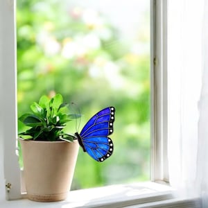 May include: A vibrant blue butterfly decoration with black trim and white dots is positioned near a small potted plant. The butterfly is affixed to a window, with a blurred green background visible through the glass.