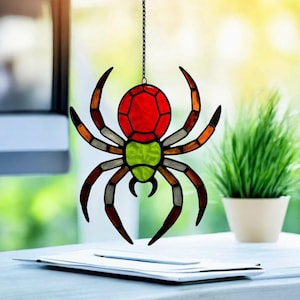 May include: A colourful stained glass spider suncatcher. The spider has a red and green body, with orange and brown legs. It hangs from a black chain. The spider is designed to catch the light.