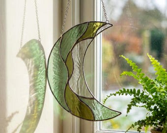 Può includere: Un acchiappasole a forma di mezzaluna in vetro colorato, nei toni del verde e del vetro trasparente, appeso a una catena d'argento. L'acchiappasole è esposto davanti a una finestra, proiettando un'ombra sul muro. Il tema è celeste e decorativo.