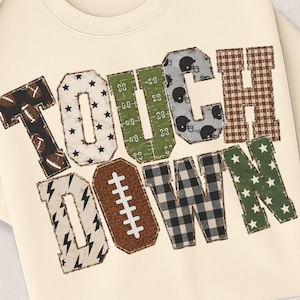 Könnte beinhalten: Ein cremefarbenes Sweatshirt mit Rundhalsausschnitt und dem Wort TOUCHDOWN in großen Blockbuchstaben. Jeder Buchstabe hat ein einzigartiges Muster, darunter Fußbälle, Sterne, ein grünes Spielfeld, Helme, braunes Karomuster und Blitze.