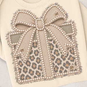 May include: A cream-colored sweatshirt featuring a large graphic of a gift box. The box has a leopard print pattern, a beige ribbon bow, and is outlined with white faux pearls. The edges of the graphic have a frayed, textured appearance.
