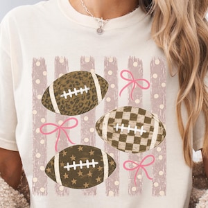 May include: A cream-colored t-shirt featuring a graphic print of three footballs with leopard, checkered, and star patterns. The design includes vertical distressed stripes, light-colored polka dots, and three pink hand-drawn style bows.