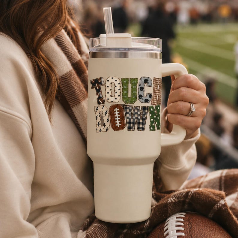 May include: A cream-colored insulated tumbler with a handle and a clear lid featuring a straw. The word TOUCHDOWN is printed in a block-letter style, with each letter filled with various football-themed patterns including brown, green, and black.