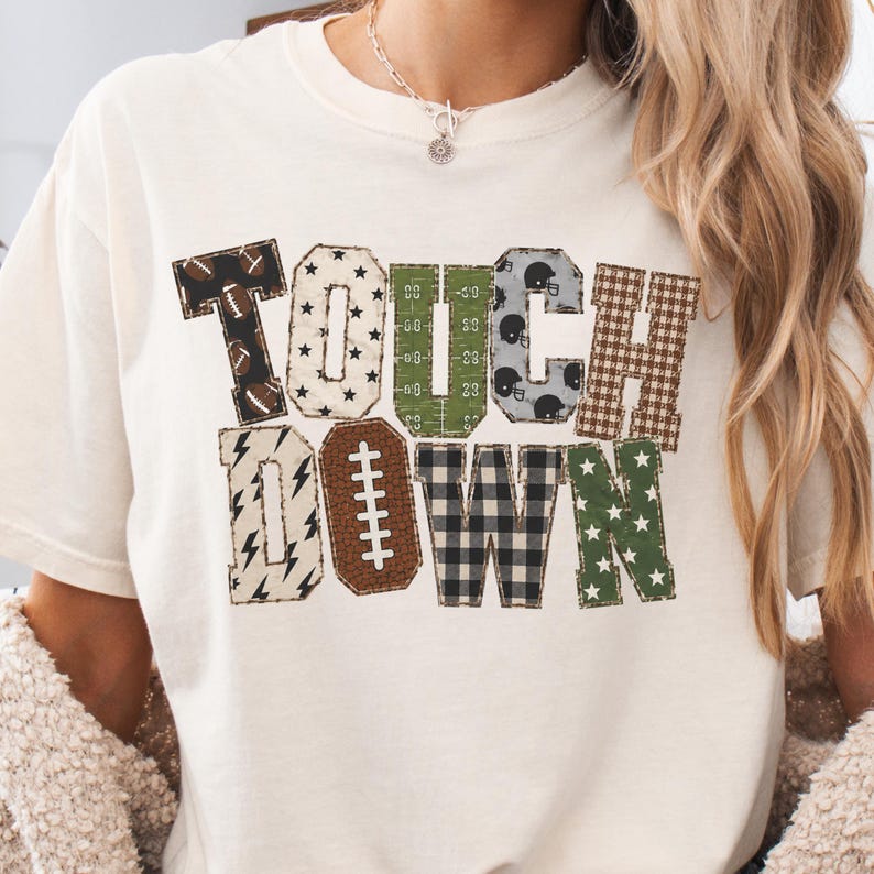 May include: A cream-colored short-sleeve t-shirt featuring the word TOUCHDOWN in large block letters. Each letter has a unique pattern including footballs, stars, field markings, helmets, plaid, and lightning bolts in brown, green, and black.