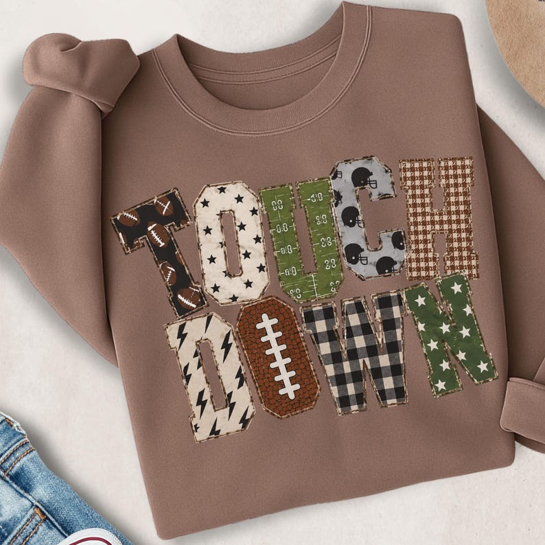 May include: A mocha brown crewneck sweatshirt featuring the word TOUCHDOWN in large block letters. Each letter has a unique pattern including footballs, stars, green field markings, black helmets, brown plaid, lightning bolts, and buffalo check.
