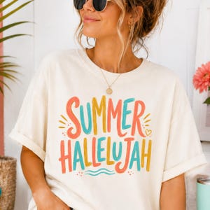 May include: An off-white short-sleeve t-shirt featuring the text "SUMMER HALLELUJAH" printed in a colorful, hand-lettered style. The graphic uses shades of coral, yellow, and teal, accented with small dots, a heart, and wavy lines.