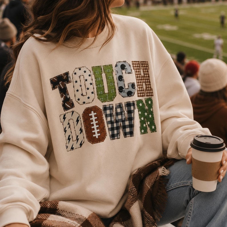 May include: A cream-colored crewneck sweatshirt featuring the word TOUCHDOWN printed in large, multi-patterned block letters. Each letter displays a unique design, including footballs, stars, plaid, and gridiron field markings in various colors.