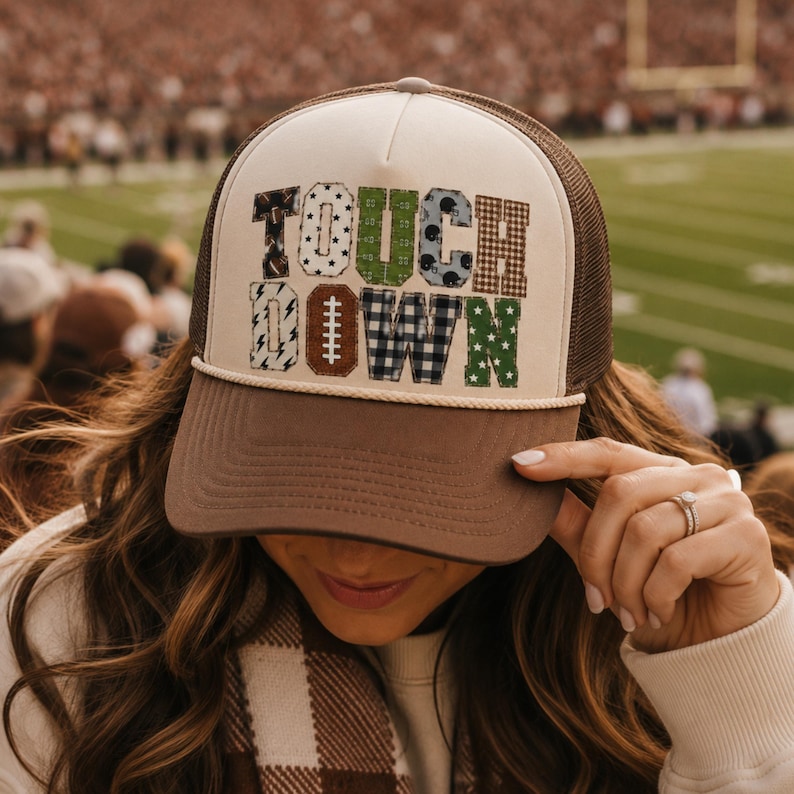 May include: A brown and cream trucker hat featuring the word TOUCHDOWN in large, block letters. Each letter has a unique pattern, including footballs, stars, plaid, and stripes. The hat has a brown brim, a cream front panel, and brown mesh back.
